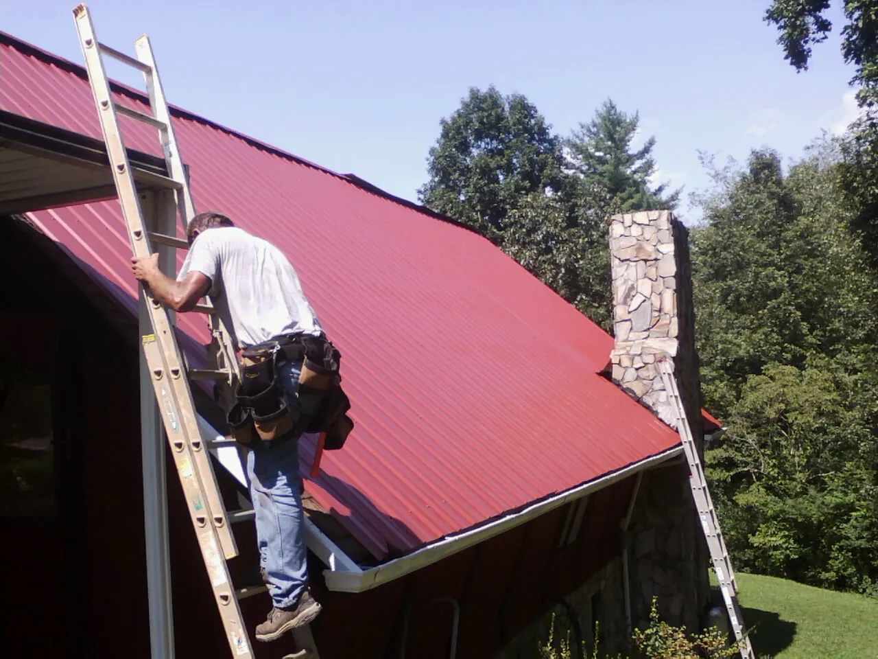 Roofer on a ladder beside a red metal roof and stone chimney — Soffit & Fascia Repair by Roofing Brothers in Valley Village